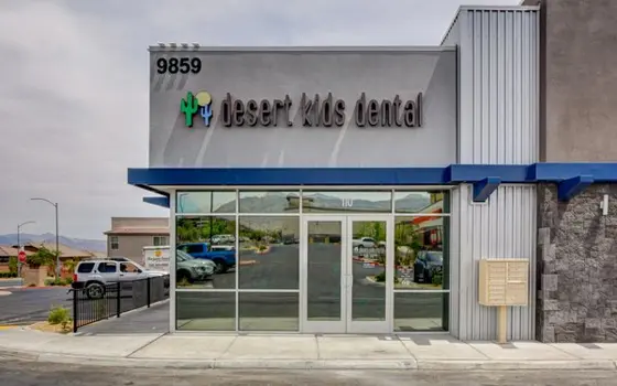 Exterior of the Desert Kids Dental clinic: a grey single-storey storefront with the clinic's name and a cactus-and-tooth logo above a blue awning and glass entrance doors, with mountains in the distance.