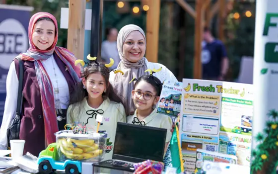 Two women in headscarves and two young students wearing banana-shaped headbands smile behind their "Fresh Tracker" project table, which holds a laptop, a box of bananas wired to sensors, and a poster explaining how the project reduces food waste.